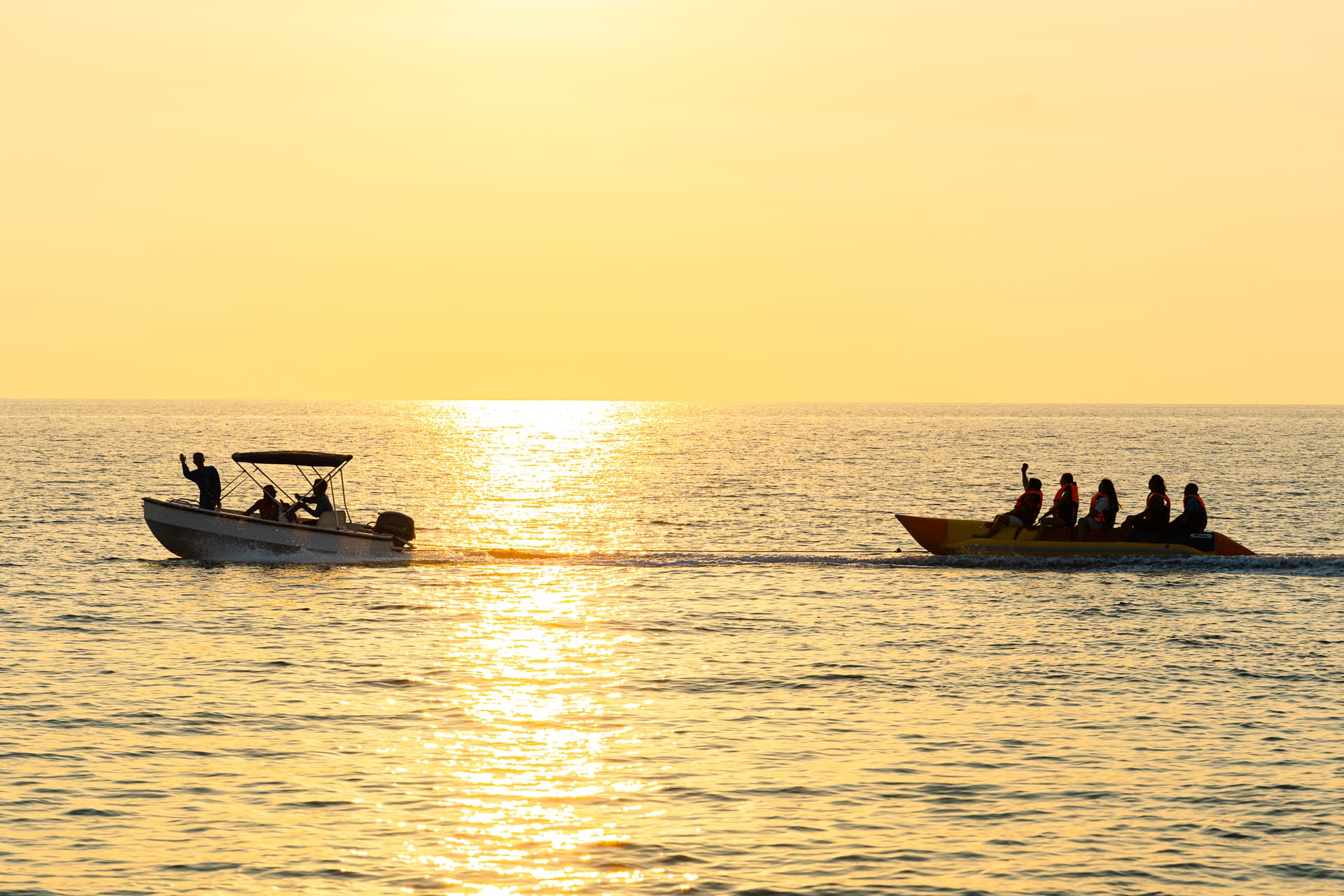 Guests on a banana-boat ride beside a motorboat on calm water at golden-hour sunset